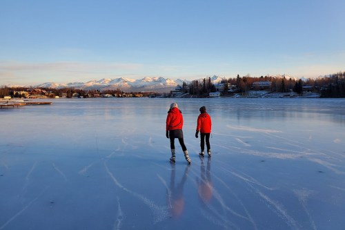 Two people in red jackets skate on a frozen lake with mountains in the background under a clear blue sky.