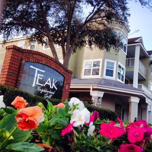 Sign for Teak Neighborhood Grill in front of a building, surrounded by colorful flowers and trees.