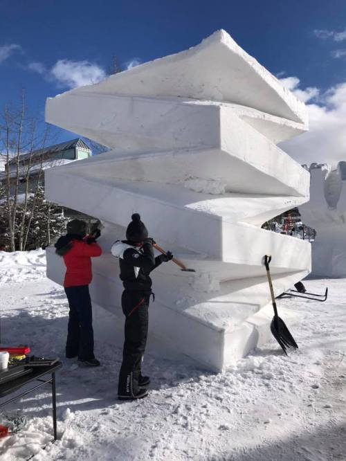 Two people sculpting a large, abstract snow structure on a sunny day, surrounded by snow and trees.