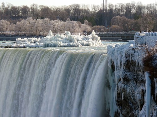 A frozen waterfall with icy formations, surrounded by snow-covered trees and a clear blue sky.