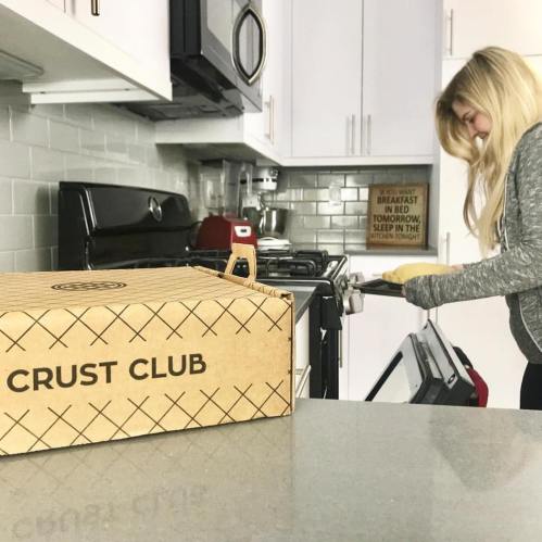 A woman in a kitchen pulls a tray from the oven, with a "Crust Club" box on the counter.
