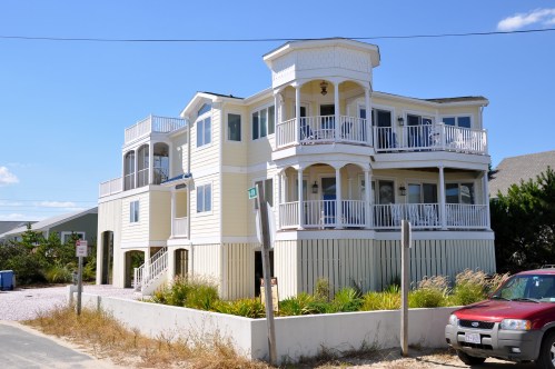 A large, multi-level beach house with balconies, surrounded by greenery and a clear blue sky.