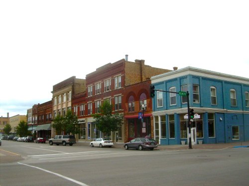 Historic buildings line a street, featuring red brick and blue facades, with trees and parked cars in view.