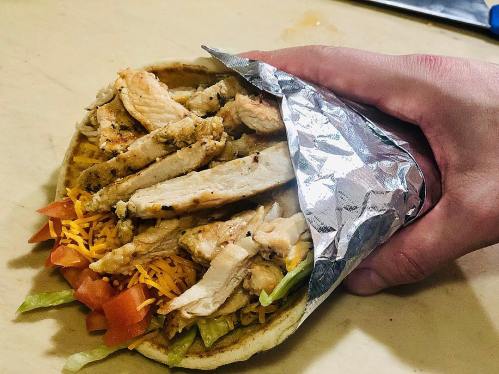 A hand holding a foil-wrapped pita filled with grilled chicken, cheese, lettuce, and tomatoes.