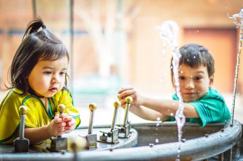 Two children play with water features, one focused and the other smiling, in a bright outdoor setting.
