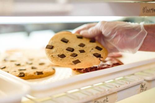 A hand in a glove lifts a chocolate chip cookie from a tray in a bakery setting.
