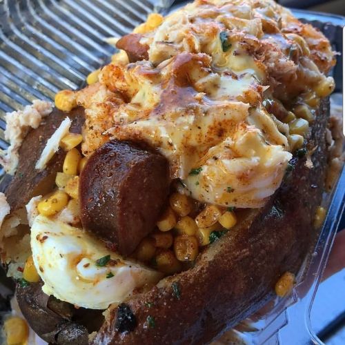A loaded baked potato topped with sausage, corn, cheese, and spices, served in a clear container.