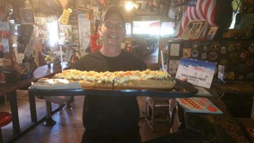 A smiling man holds a large hot dog topped with lettuce and other condiments in a lively restaurant setting.