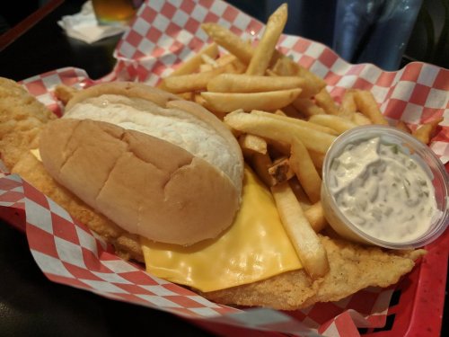 A plate with a fish sandwich on a bun, melted cheese, fries, and a side of dipping sauce in a red-checkered basket.