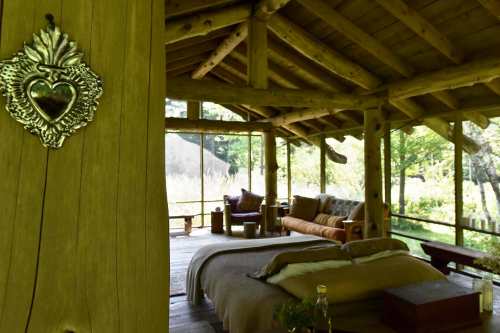 Cozy wooden cabin interior with a bed, rustic decor, and large windows overlooking nature.