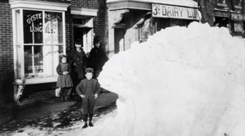 A historic black-and-white photo of children and adults outside a shop, with a large snowbank in the foreground.