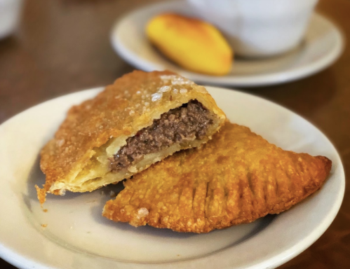 A close-up of a golden-brown empanada, partially sliced to reveal a savory beef filling, on a white plate.