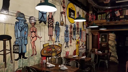 Colorful mural on a restaurant wall featuring cartoon figures and a cozy dining area with wooden tables and metal chairs.