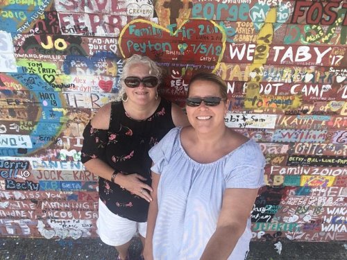 Two women pose for a selfie in front of a colorful wall covered in names and messages. Both are wearing sunglasses.