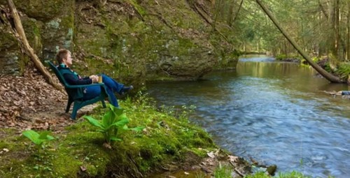 A person sits in a chair by a serene river, surrounded by lush greenery and rocky cliffs.