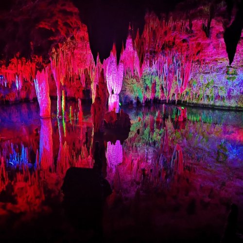 Colorful stalactites and stalagmites reflected in water within a cave, illuminated by vibrant lights.