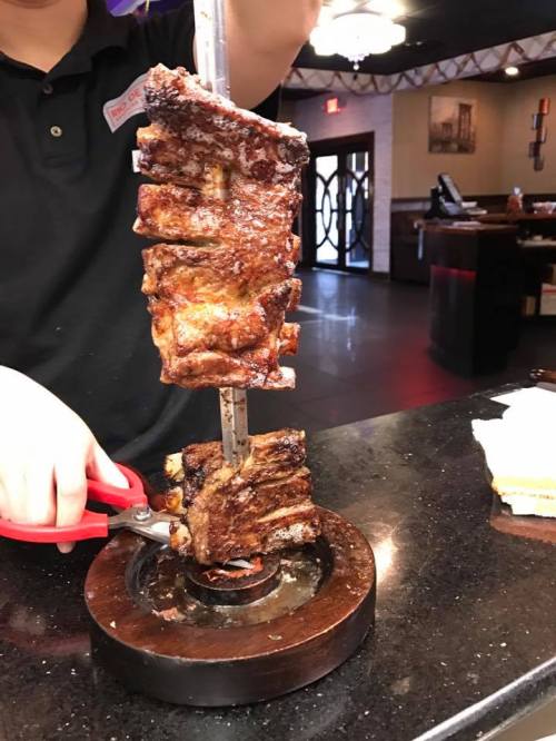 A person holds a skewer of grilled meat, ready to slice portions at a restaurant.