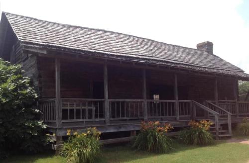A rustic wooden house with a sloped roof, surrounded by greenery and flowers, featuring a porch and steps.