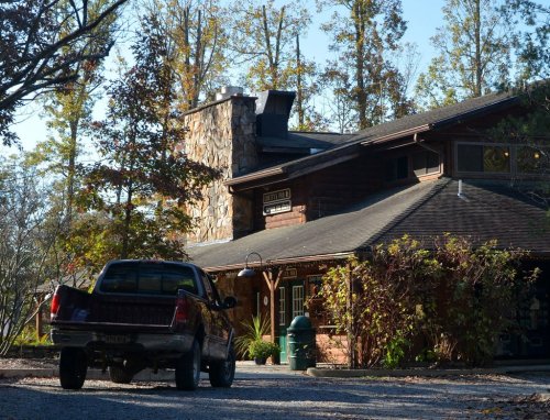 A rustic lodge with a stone chimney surrounded by trees, featuring a parked truck in the foreground.