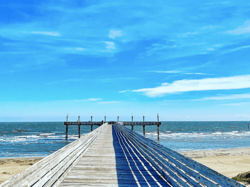 A wooden pier extends into the ocean under a bright blue sky with scattered clouds. Waves gently lap at the shore.