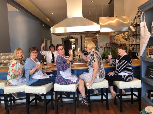 A group of six women in aprons sit at a bar, smiling and holding drinks in a bright, modern kitchen setting.