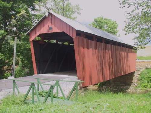 A red covered bridge with a wooden structure, surrounded by greenery and a gravel path leading to it.