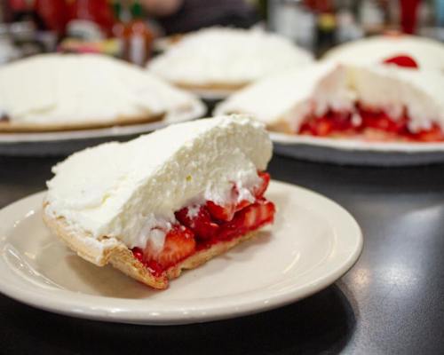 A slice of strawberry pie with whipped cream on a plate, surrounded by whole pies in the background.