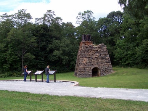 Two people stand near a stone structure in a park, surrounded by trees, with informational signs nearby.