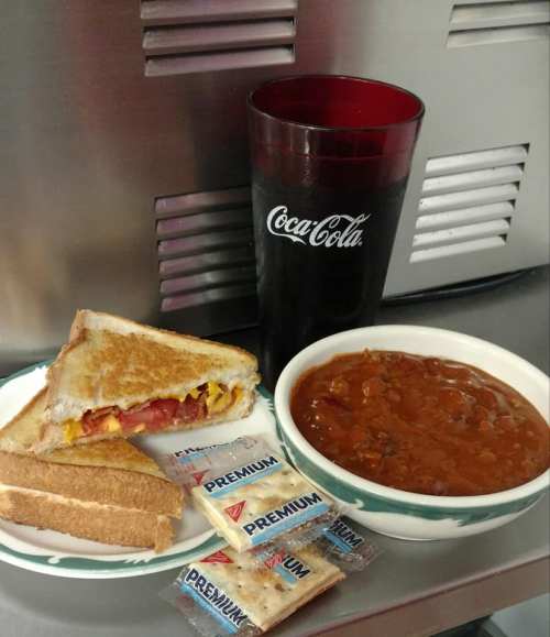 A plate with a grilled cheese sandwich, a bowl of chili, and crackers, alongside a glass of Coca-Cola.