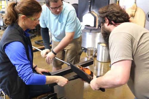 A glassblowing workshop scene with three people working together on a glass piece. Safety gear is worn.