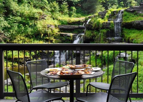A table with food set on a balcony overlooking a lush green landscape and a waterfall.