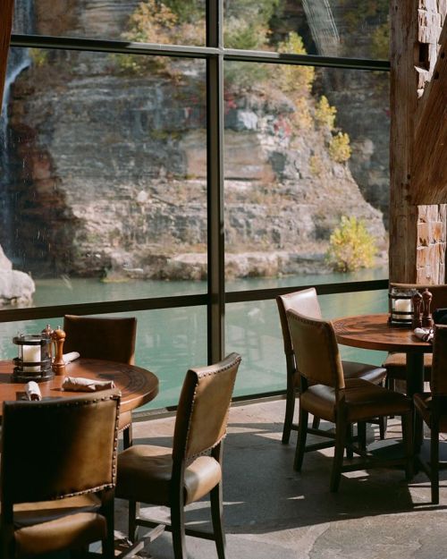 Cozy restaurant interior with wooden tables and chairs, overlooking a scenic view of a river and rocky cliffs.