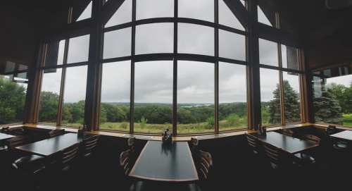 A spacious restaurant with large windows offering a view of green trees and a cloudy sky. Tables are set for dining.