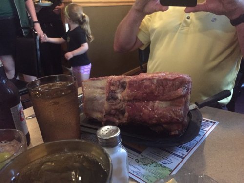 A large piece of meat on a sizzling plate at a restaurant, with drinks and a person in a yellow shirt nearby.
