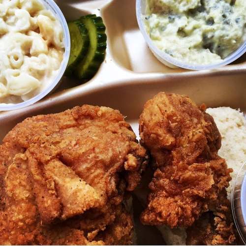 A takeout tray with fried chicken, macaroni and cheese, pickles, and a side of green sauce.