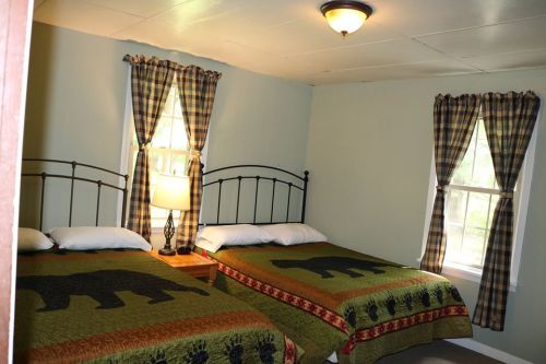 Cozy bedroom with two beds, bear-themed quilts, and plaid curtains, featuring a lamp and natural light from windows.