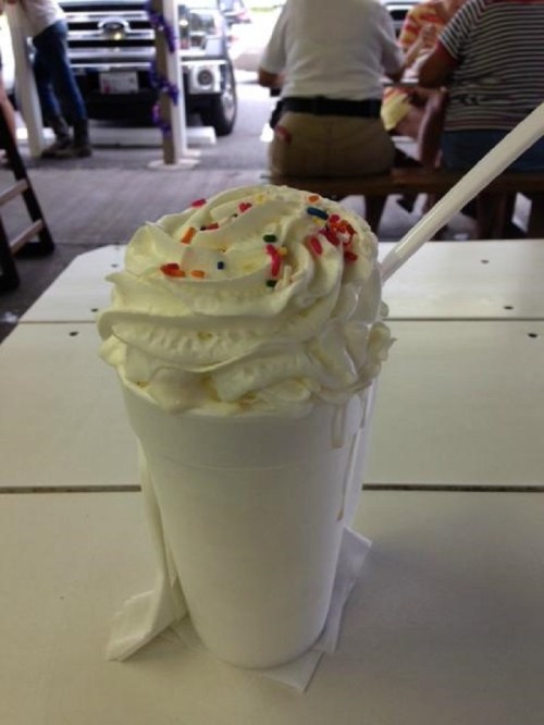 A white cup filled with whipped cream and colorful sprinkles, placed on a table with people in the background.