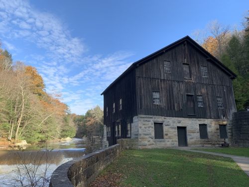 Historic wooden mill beside a river, surrounded by trees and a clear blue sky. Autumn foliage is visible.