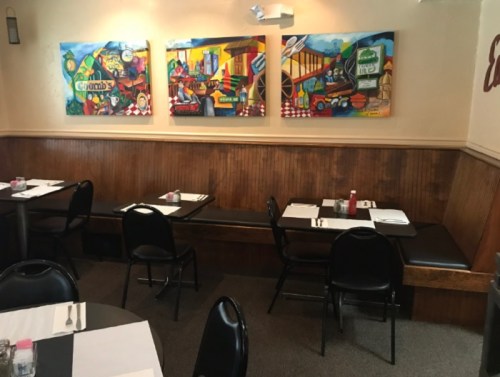Interior of a restaurant featuring colorful artwork on the walls and empty tables with black chairs.