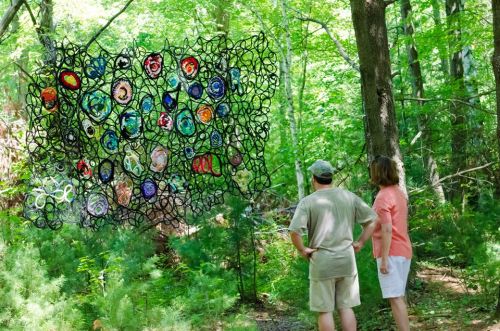 Two people admire a colorful, abstract art installation in a lush green forest.