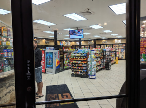 A convenience store interior with shelves of snacks and drinks, a TV screen, and a customer near the entrance.