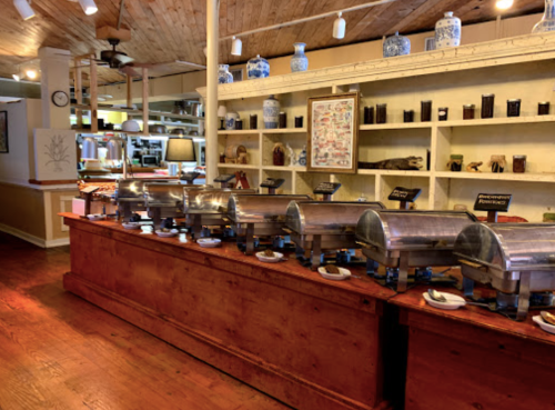 A buffet setup with metal chafing dishes on a wooden counter, surrounded by shelves of decorative items and a cozy interior.