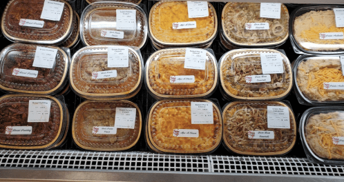 A display of various prepared meals in clear containers, labeled with names and prices, arranged in a refrigerated case.