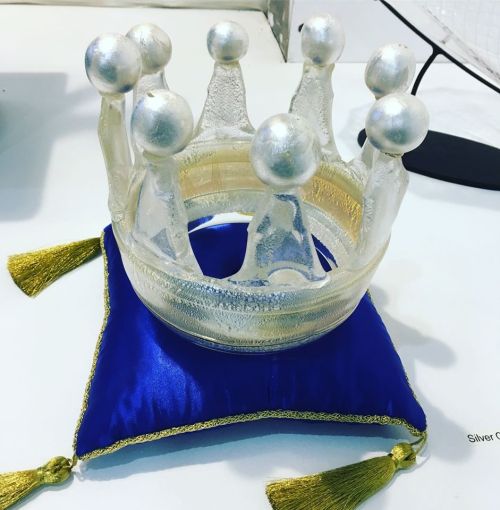 A glass crown with silver orbs sits on a blue pillow with gold tassels.