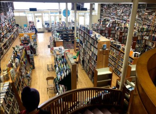 A spacious library with tall bookshelves, wooden floors, and large windows, filled with books and a few chairs.