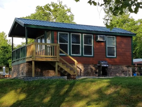 A small cabin with a wooden deck, stone foundation, and green trim, surrounded by trees and grass.