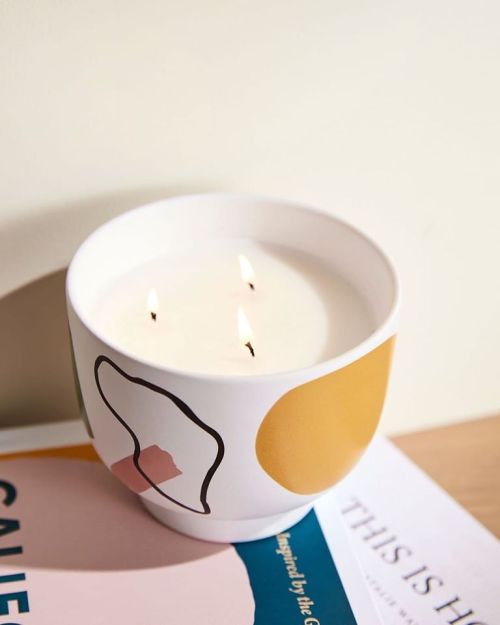 A decorative candle with three wicks sits on a stack of magazines, featuring abstract designs in pastel colors.