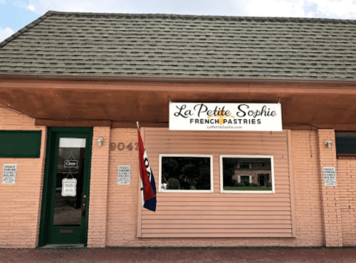 Exterior of La Petite Sophie, a French pastries shop, featuring a sign and a flag, with a brick facade and windows.