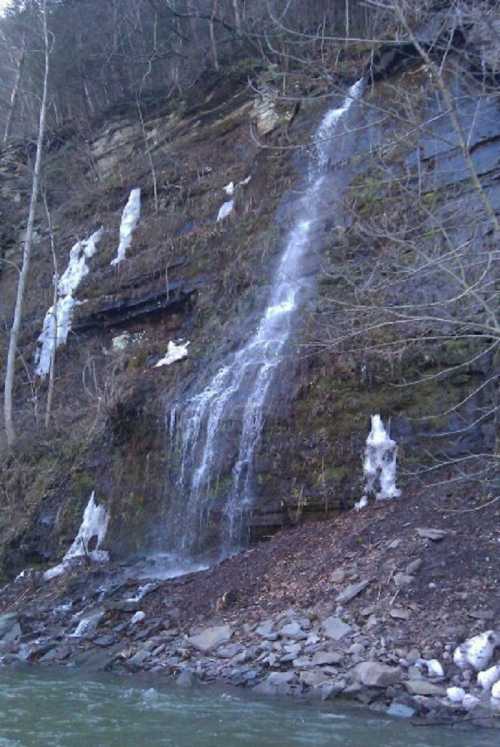 A waterfall cascading down a rocky cliff, surrounded by trees and patches of ice along the water's edge.