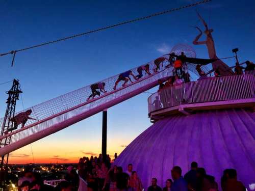 People climbing a net structure at sunset, with a city skyline in the background and a dome-shaped building below.